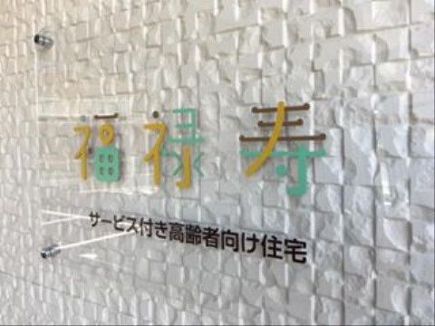 白い凸凹のある壁に、施設名を書いたプレートが設置されている。３色で書かれた文字がおしゃれで、さわやかさを感じる。