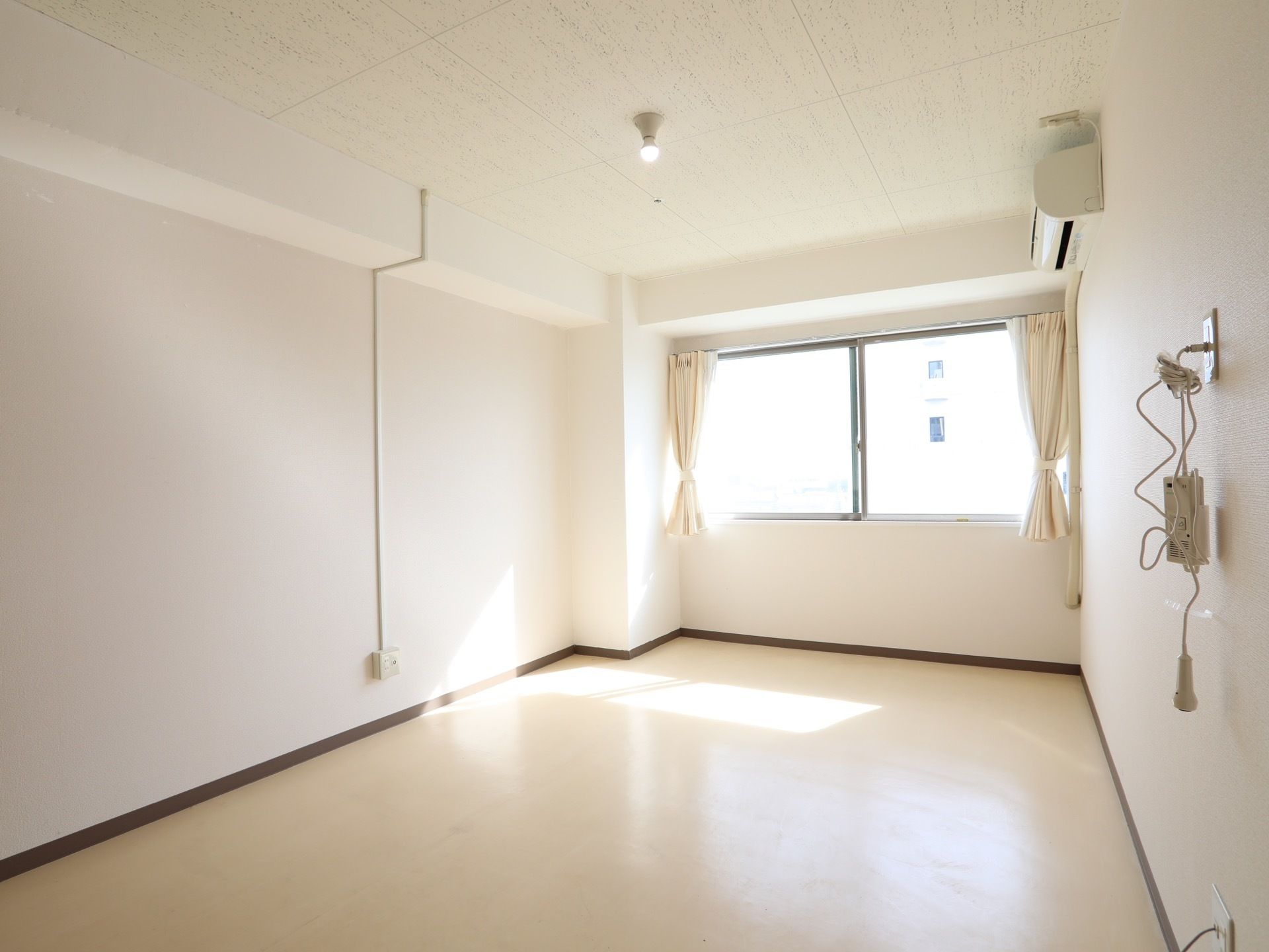 白を基調とした明るい居室である。窓にはカーテンが取り付けてあり、壁にはエアコンが１台とコンセントの差し込み口が設置されている。