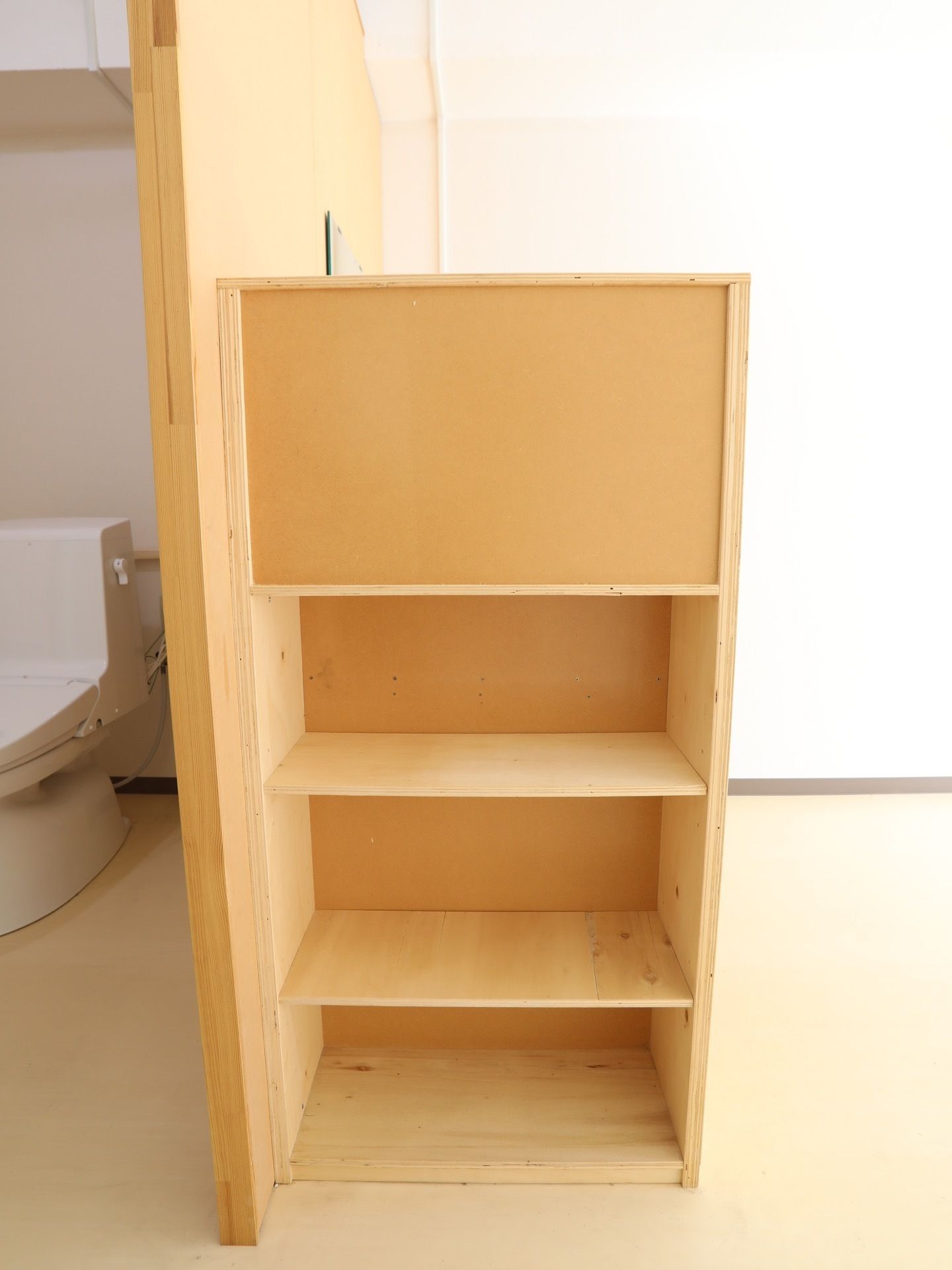 白い壁の居室である。木製の３段に仕切られた棚が床に置かれている。奥にはトイレがあり、扉は開いている。