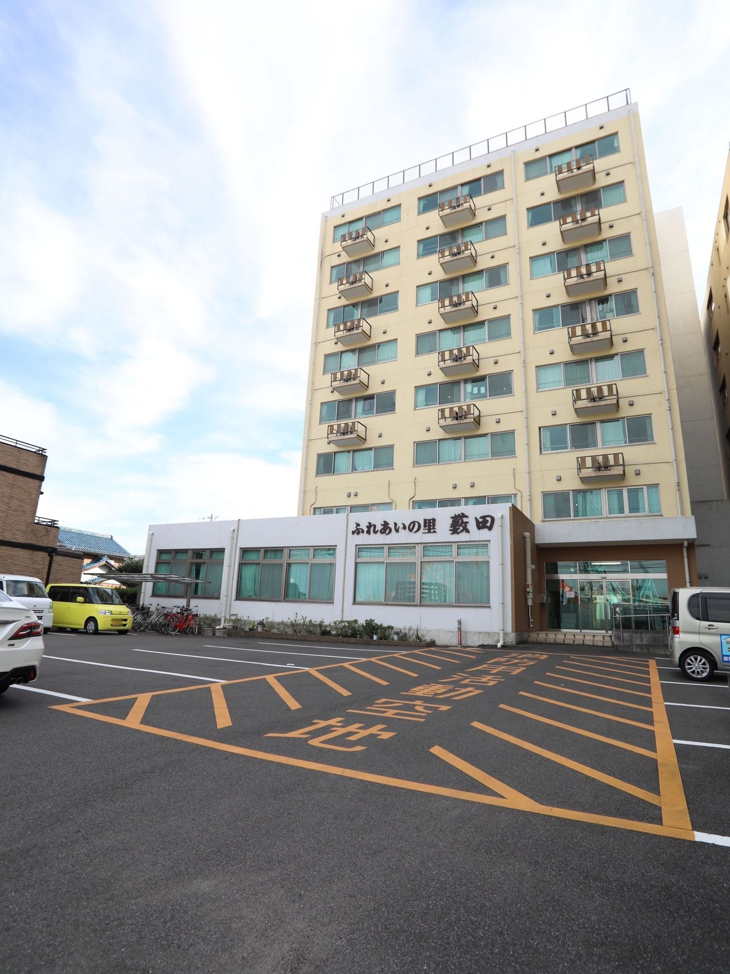 ８階建てのコンクリート造りの建物である。前には、駐車スペースが確保されており、車が数台停まっている。