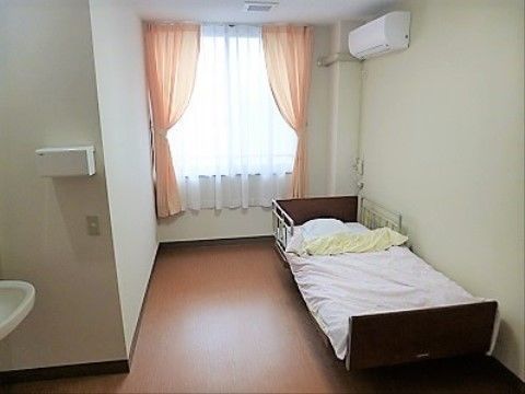 逆Ｌ字型の部屋にベッドが置いてあり、車椅子対応の洗面台が設置されている。腰高窓にピンク色のカーテンとレースカーテンが掛けられている。