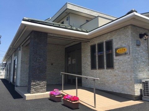 大きな一戸建のような２階建ての建物。入口にはスロープが設置されていて、その横には色鮮やかな花が咲いたプランターが置かれている。
