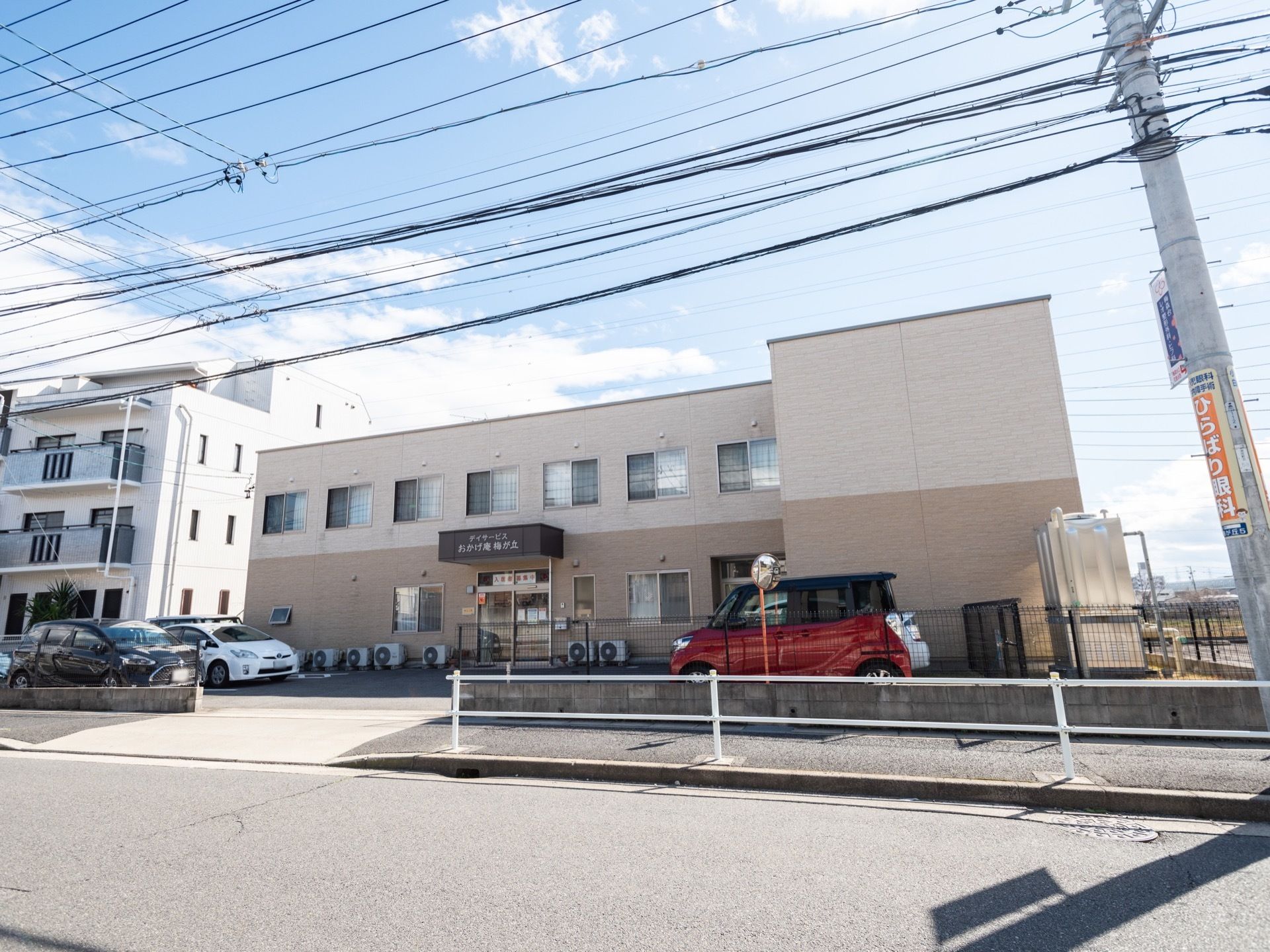 薄いベージュと濃いベージュの落ち着いた雰囲気の建物である。建物の前には、施設専用の駐車スペースがある。