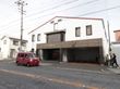 施設の建物が、歩道沿いに建っている。2階建てで、1階の右側は駐車スペースになっている。左側には、玄関がある。
