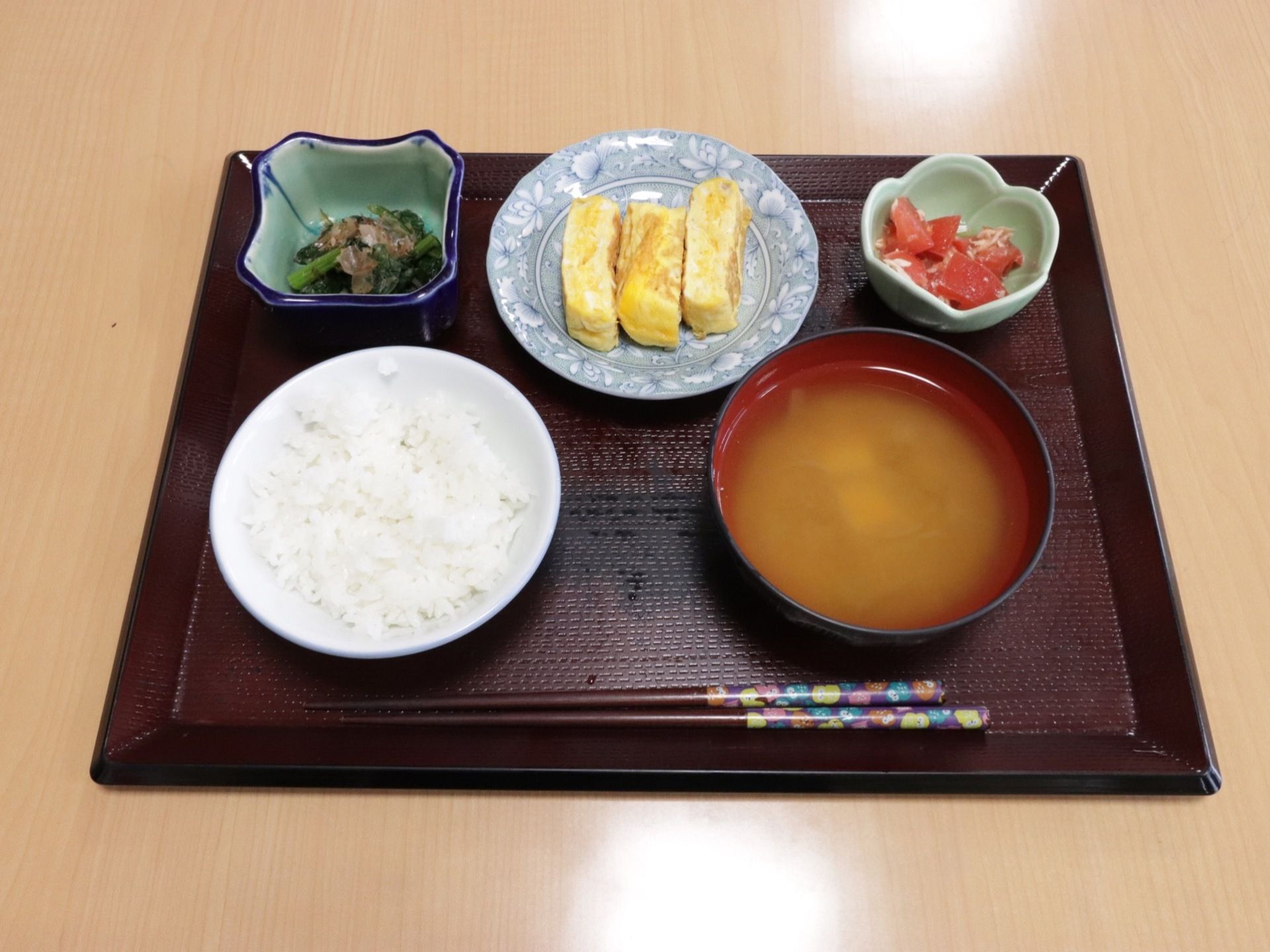 木のトレイに、ご飯とみそ汁、卵焼き、ほうれん草のお浸し、トマトが入った小鉢が並べられている。お箸も置かれている。