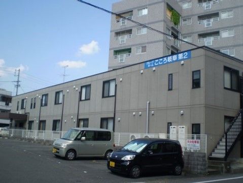 外壁が濃いベージュ色の２階建の建物は、隣の敷地との間に白く細いフェンスで仕切られている。玄関上には濃い茶色のひさしがある。