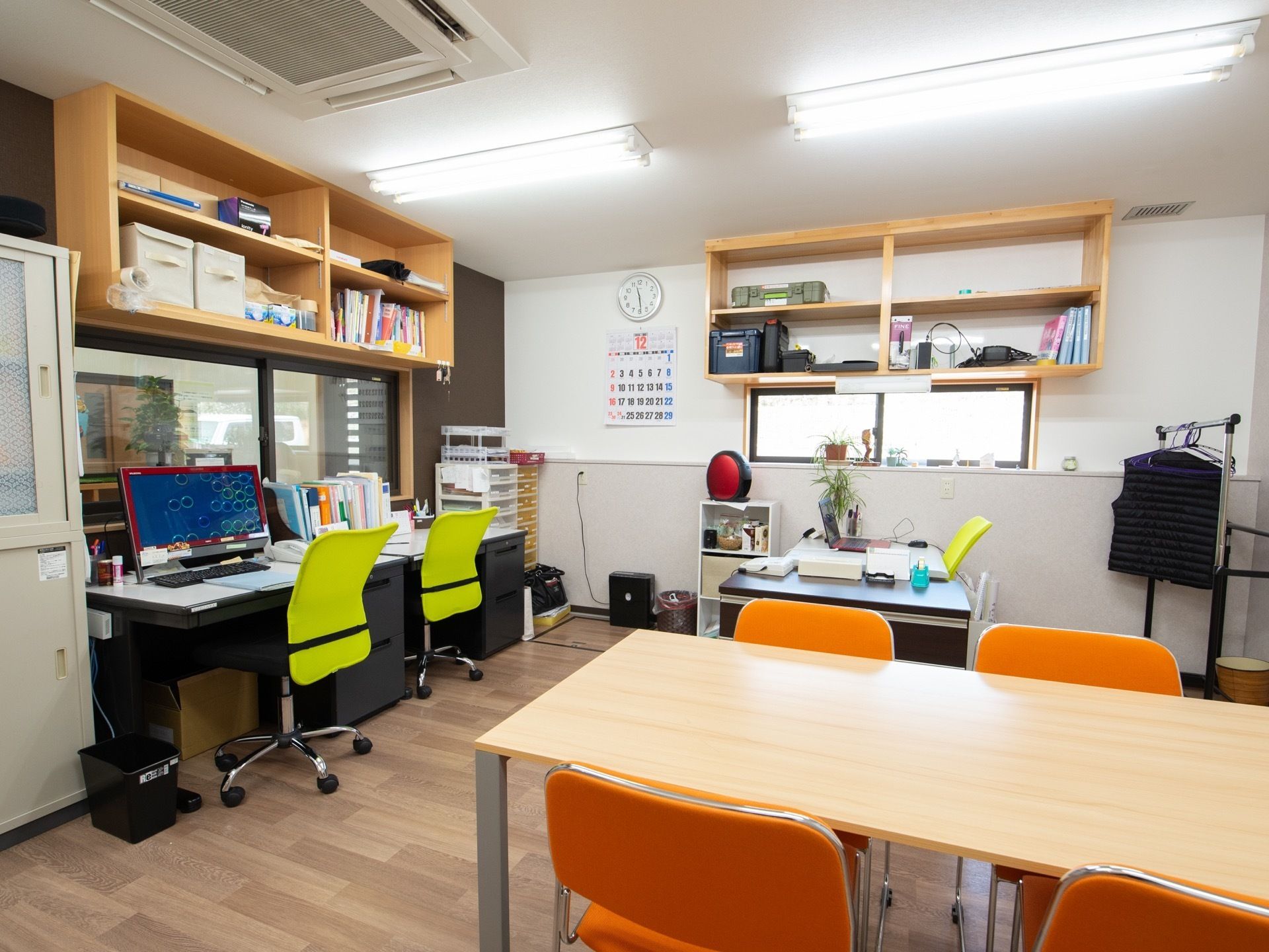 事務室の机の上に、パソコンのモニターが置かれている。打ち合わせ用のテーブルと椅子が部屋の中央部にある。