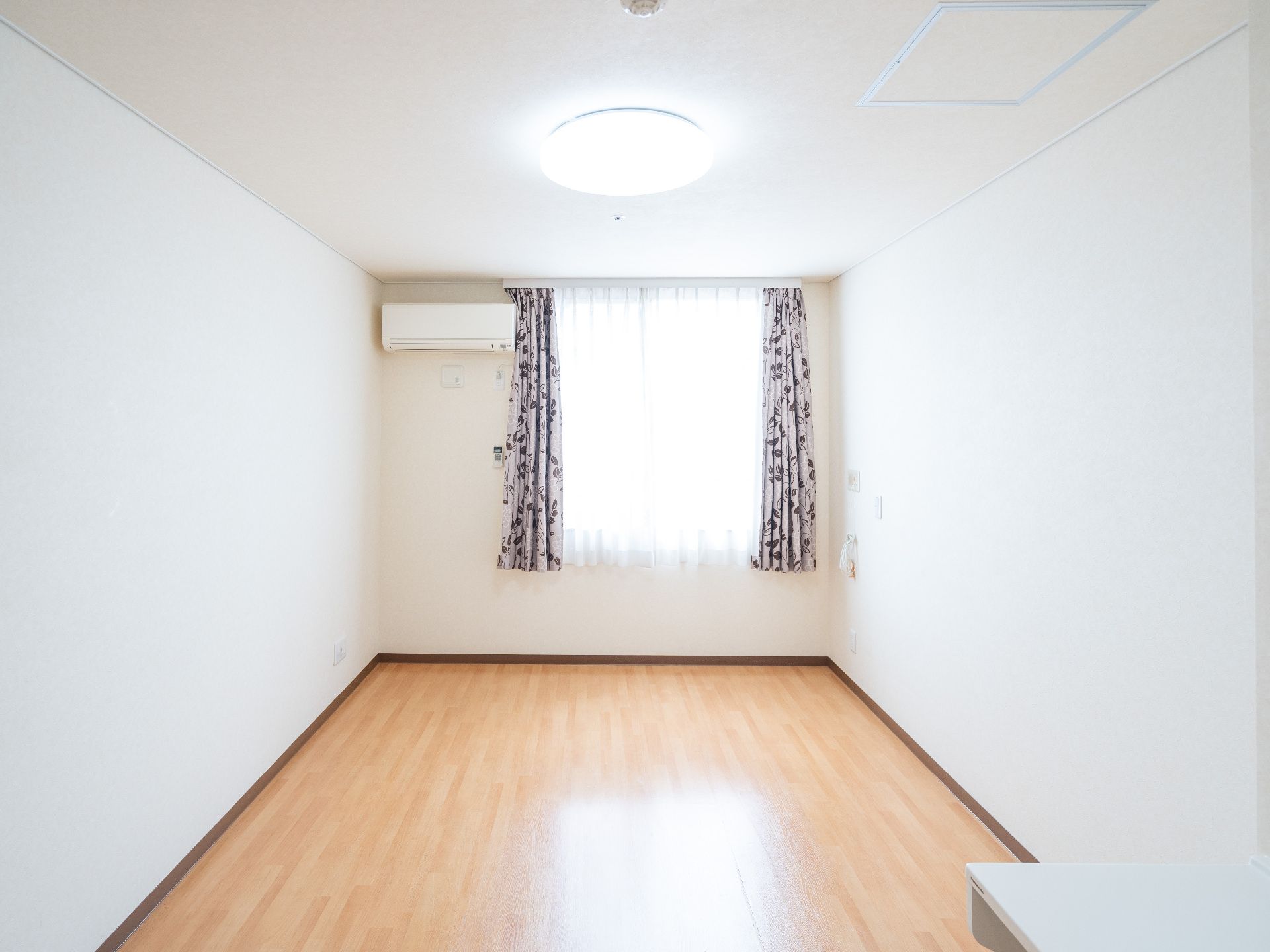 壁にカーテンのついた窓とエアコンがある何も置かれていない白い部屋