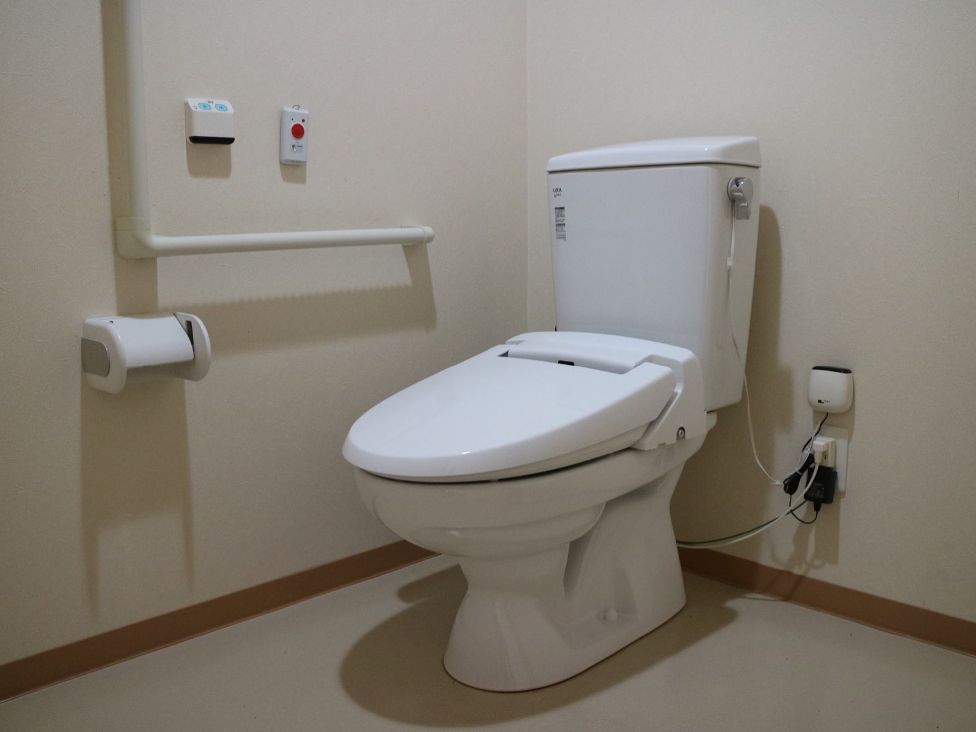 居室に完備されたトイレである。Ｌ型手すりや呼び出しボタン、便器洗浄ユニットのリモコンが壁に設置されている。