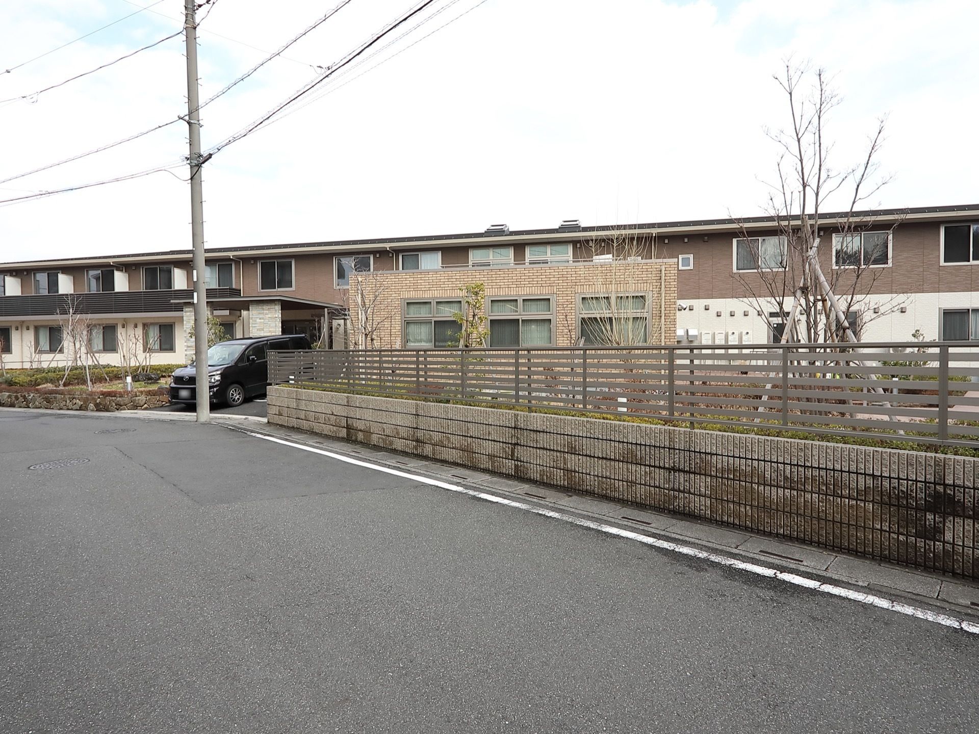道路から見ると庭の向こう側に、２階建ての建物がある。１階は白い壁で２階は茶色の壁に白い枠の窓がついている。