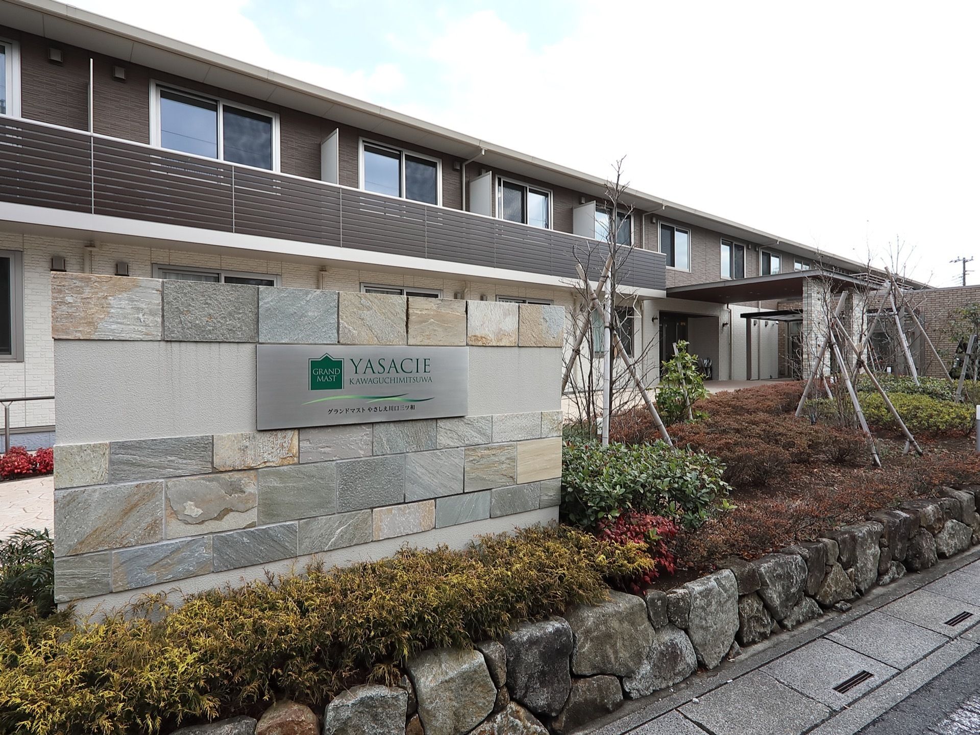 施設の建物は２階建てで、上階にはベランダを設けている。建物前にはさまざまな植物を植え、施設名を書いたプレートを設置している。