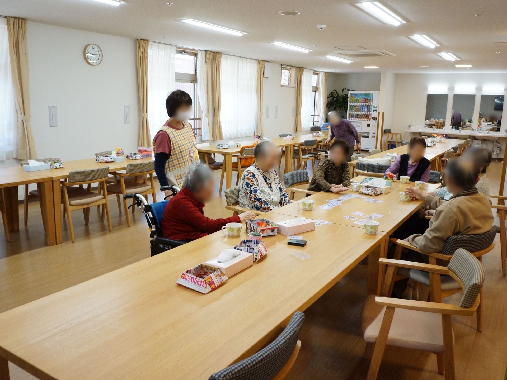 食堂兼共有スペースである。広い空間にテーブルと椅子が置かれ、数人の入居者様とスタッフが遊んだり談笑をしている。