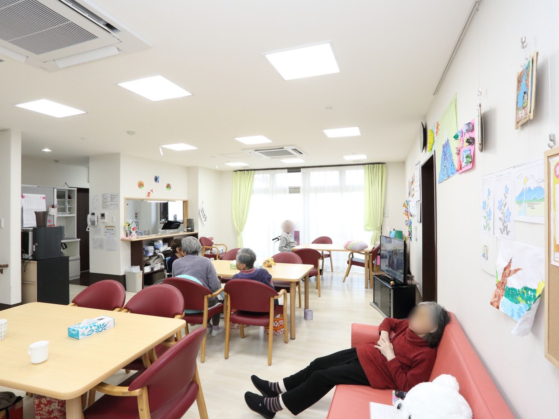 カウンターキッチンがある。明るい空間となっている。テーブル、椅子、テレビがある。壁に手作り品が飾られている。