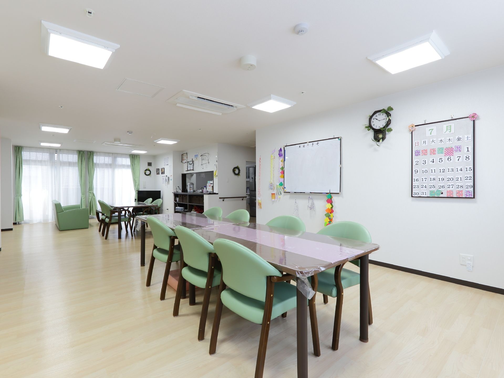 壁にカレンダーと白板がかかっている。四人掛けのテーブルが三台と、ソファーセットがある。奥に給湯用のカウンターがある。