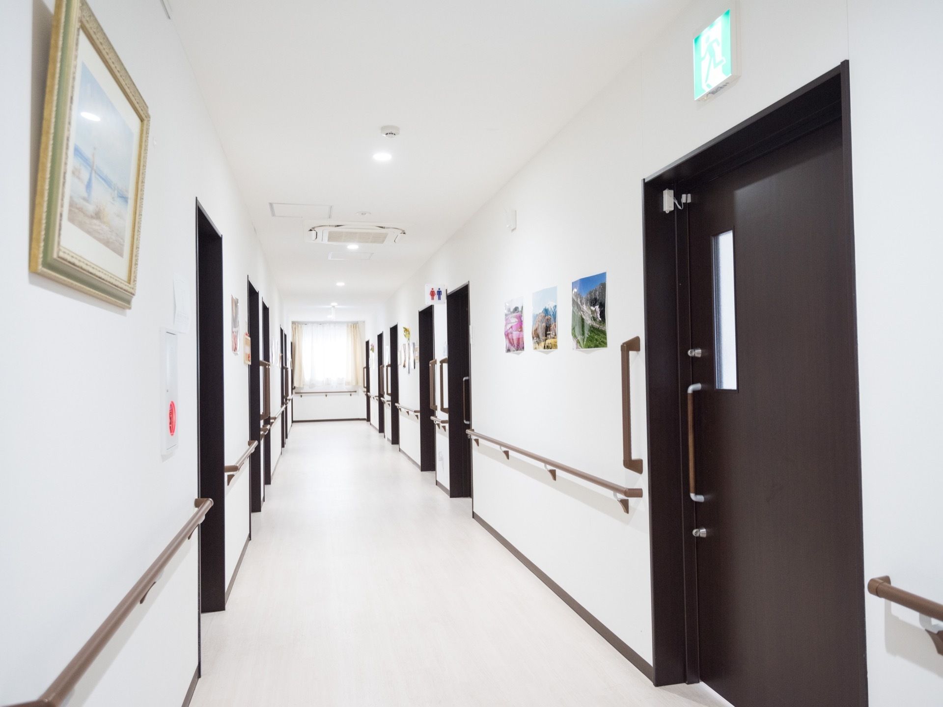 白い壁の間に、落ち着いた色合いの居室ドアが並んでいる。壁には、複数の絵が飾られているほか、手すりが設置されている。