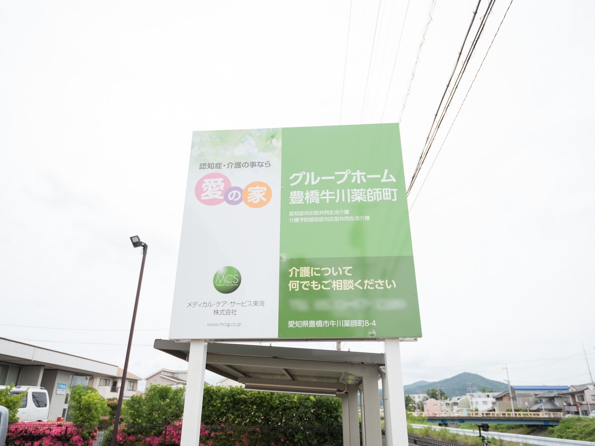 施設名が書かれた四角い看板が立てられている。周辺は豊かな自然に恵まれており、奥に橋が架かっているのが見える。