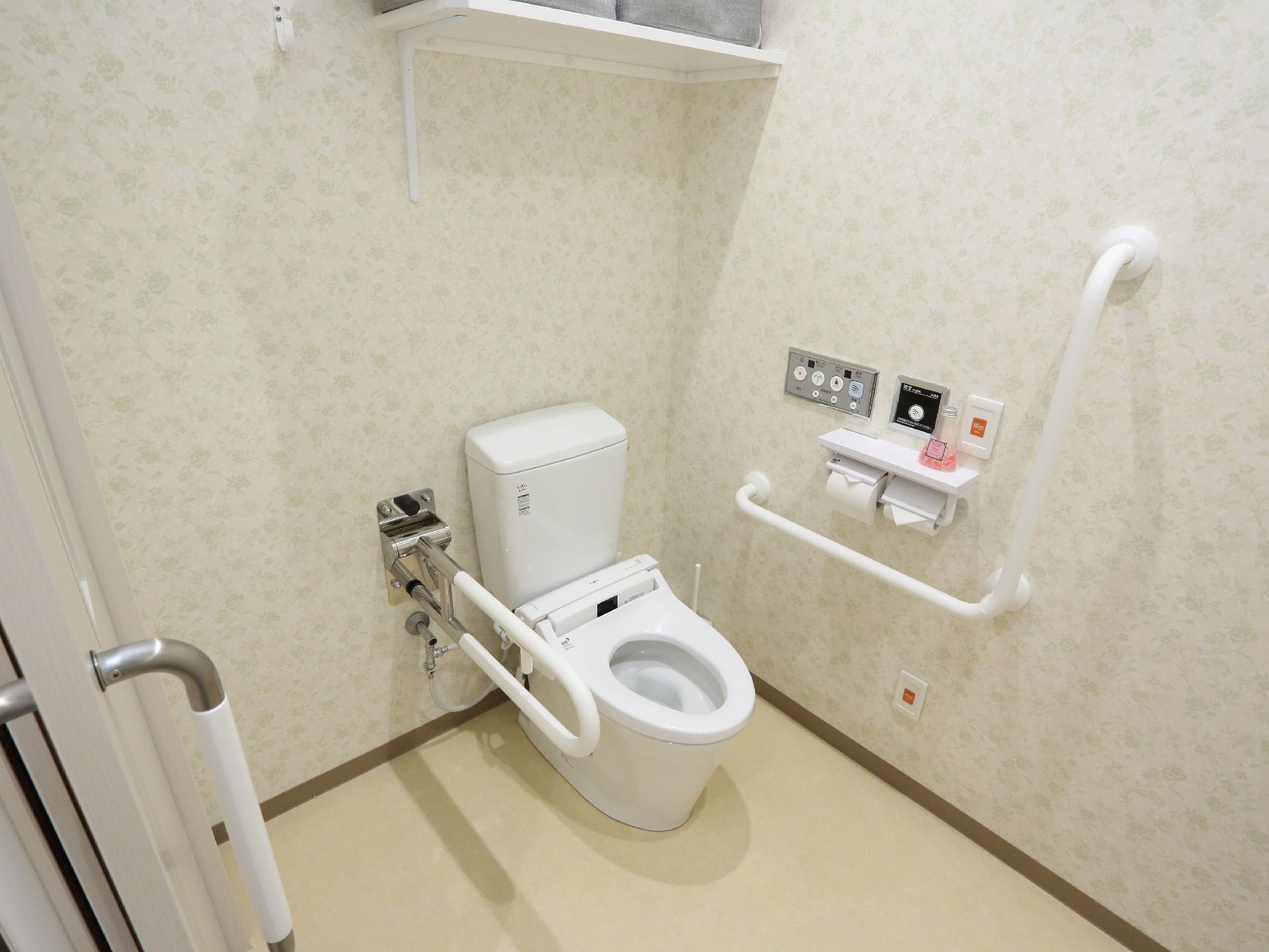 右側の壁にはL字型の手入れと呼び出しベルとティッシュペーパーがある。便器を挟んで反対側には可動式の手すりがある。