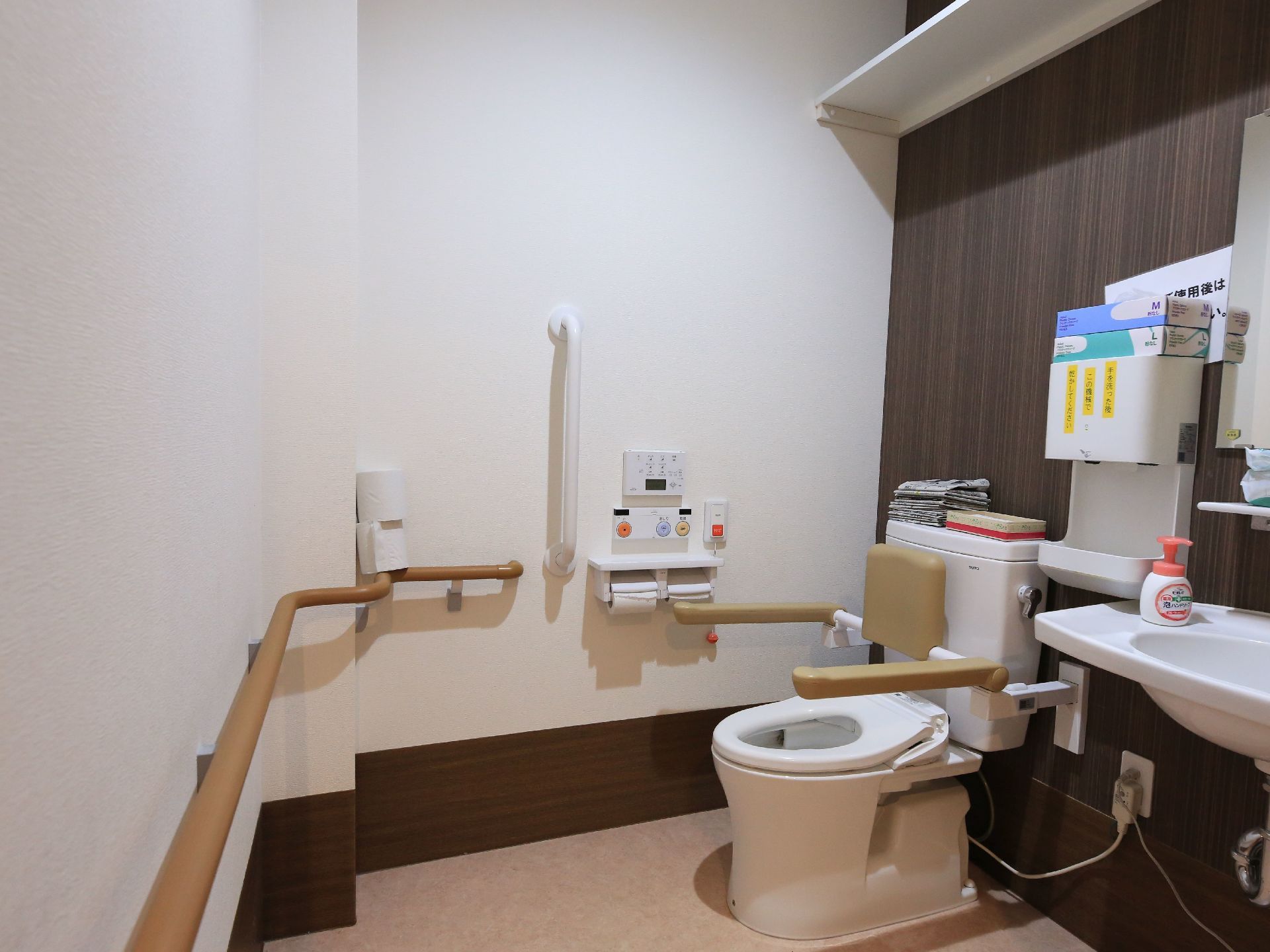 洗浄暖房便座のトイレがある。背もたれやひじ掛けがあり、壁面には大きな手すりが設置されている。ナースコールボタンもある。