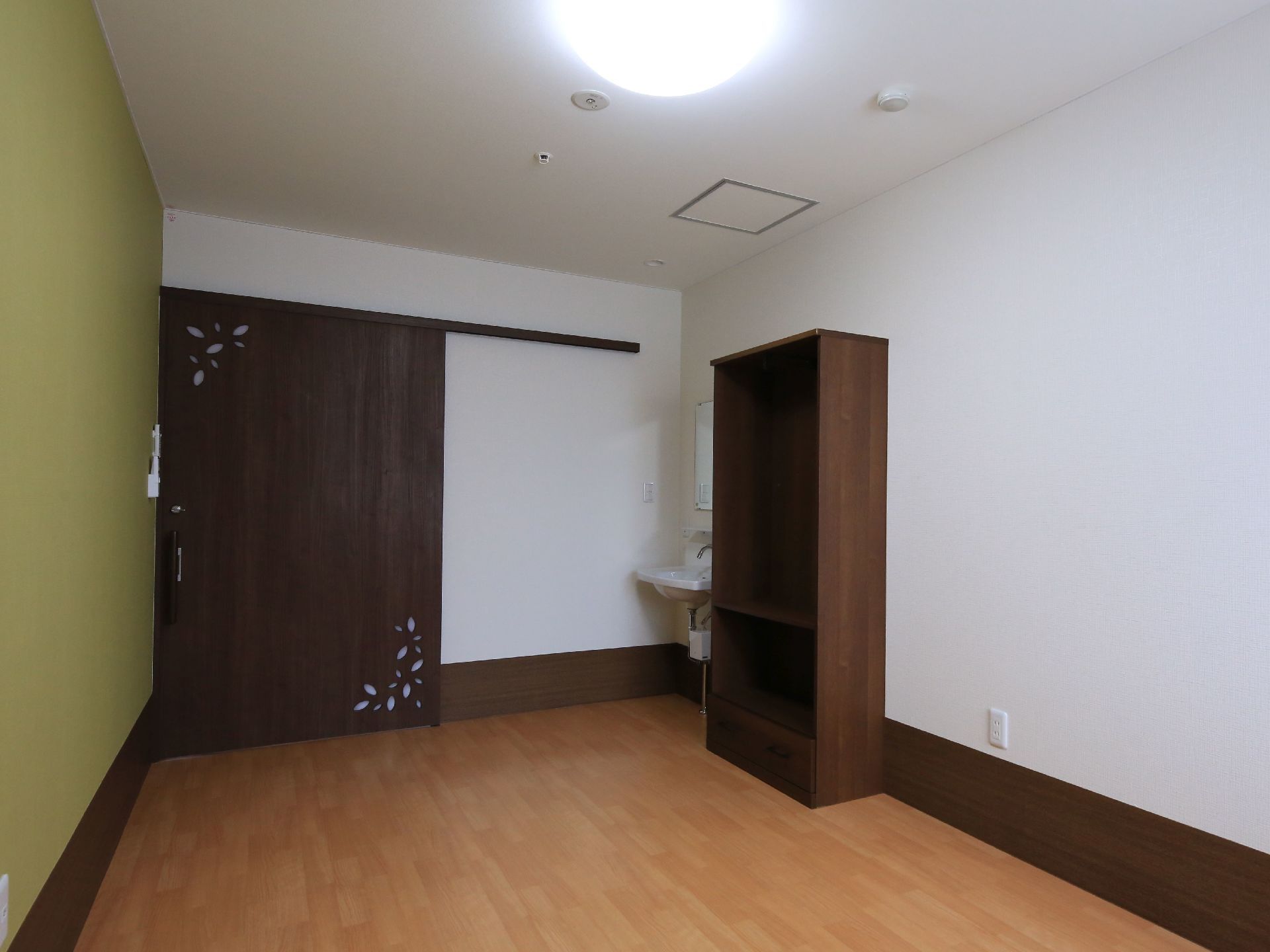 居室の入口は濃い茶色の幅の広い引き戸になっている。扉とほぼ同じ色の収納棚が洗面台のすぐ横に設置されている。