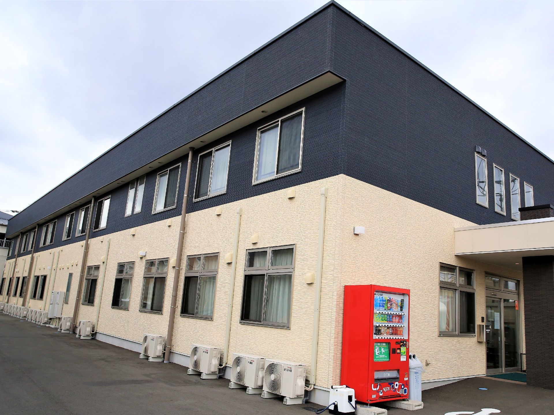１階部分は白色、２階は黒系統の色の外壁になっている施設である。玄関脇には、飲料の自動販売機が設置されている。