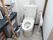 共用の個室トイレの内部。掃除用具や汚物入れなどが置かれている。便座にはひじ掛け兼手すりが取り付けてある。