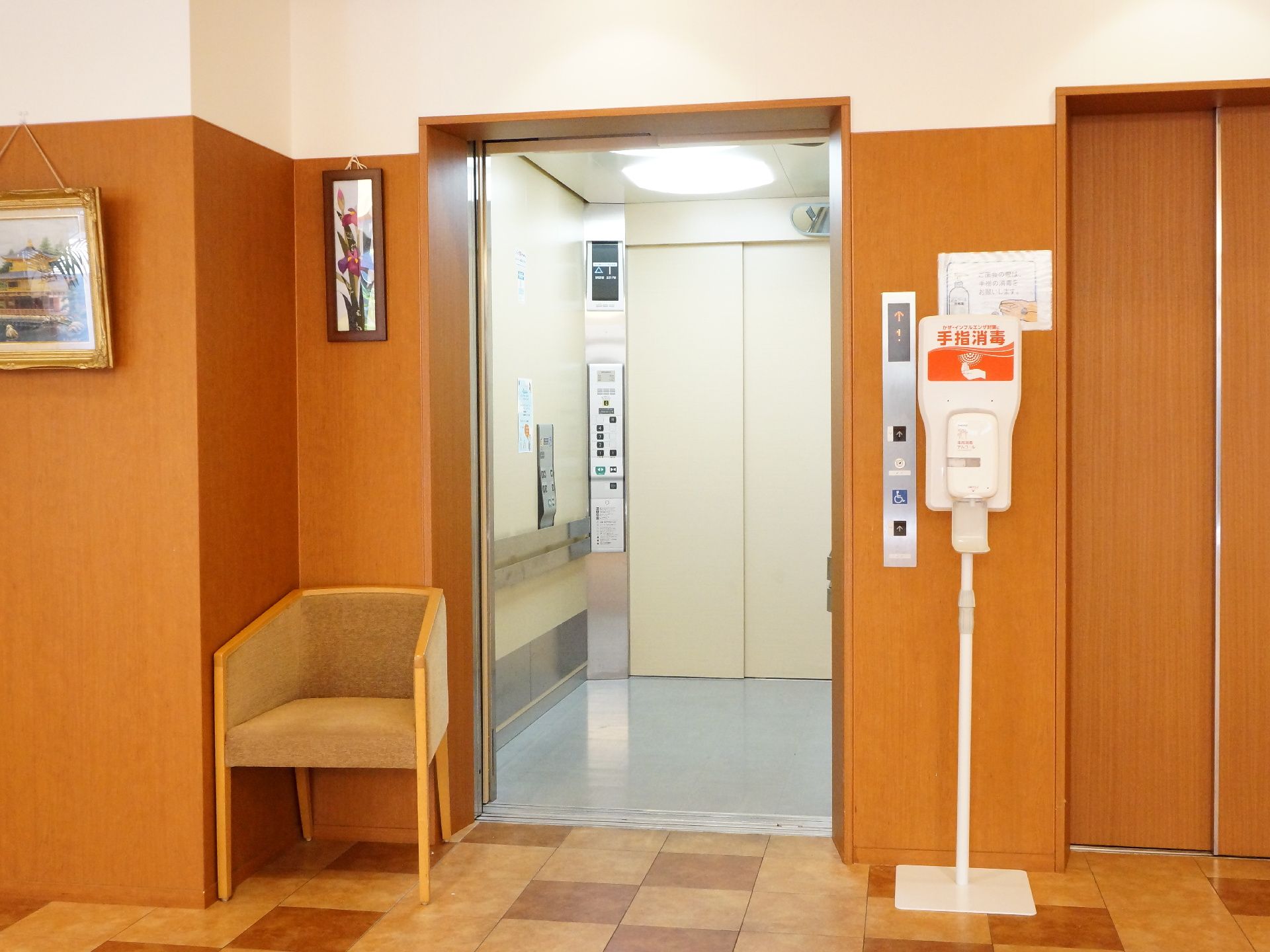 エレベーターがあるホールに消毒液と椅子が設置されている。エレベーター内部は明るく、手すりが設置されている。