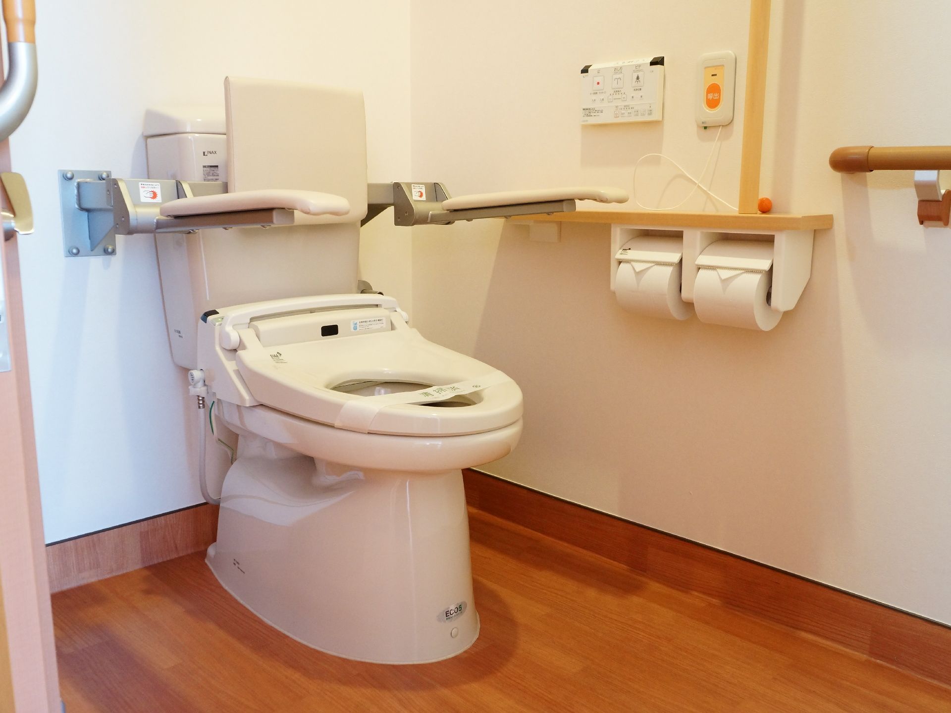 白壁に木目の床のトイレである。便器には背もたれ、両側にはひじ掛けがある。壁際には手すりとナースコールが取り付けてある。
