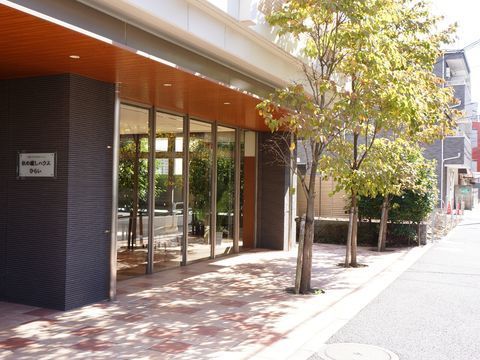 施設の敷地は歩道に面している。歩道との境には木が3本植えられている。入り口には施設名の看板がある。1階はガラス張りの個所がある。