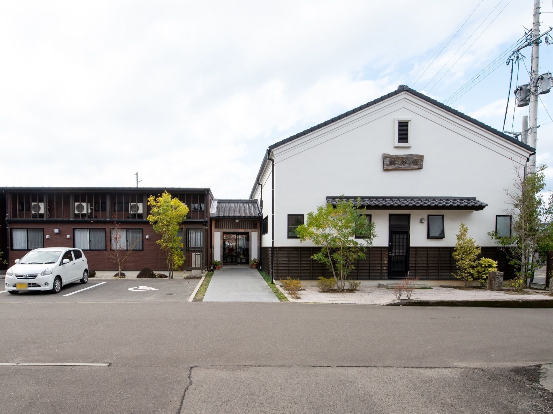 施設は、白色と茶色で統一された2階建ての建物となっている。左側には駐車スペースがあり、車が1台停められている。