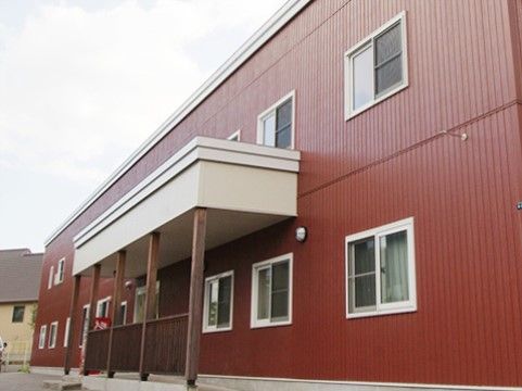 赤茶色の２階まである施設には、白い枠の窓が付いている。玄関前の白い屋根には、木製の柱と柵が設置してある。