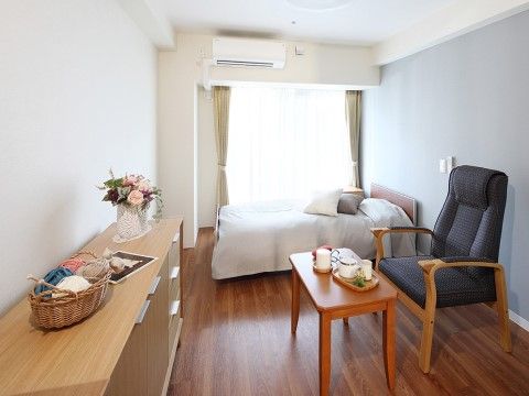 居室の窓からは外光が差し込んでおり、室内にはベッド、テーブル、手すり付きの椅子が置かれている。壁際の収納家具の上には花が飾られている。