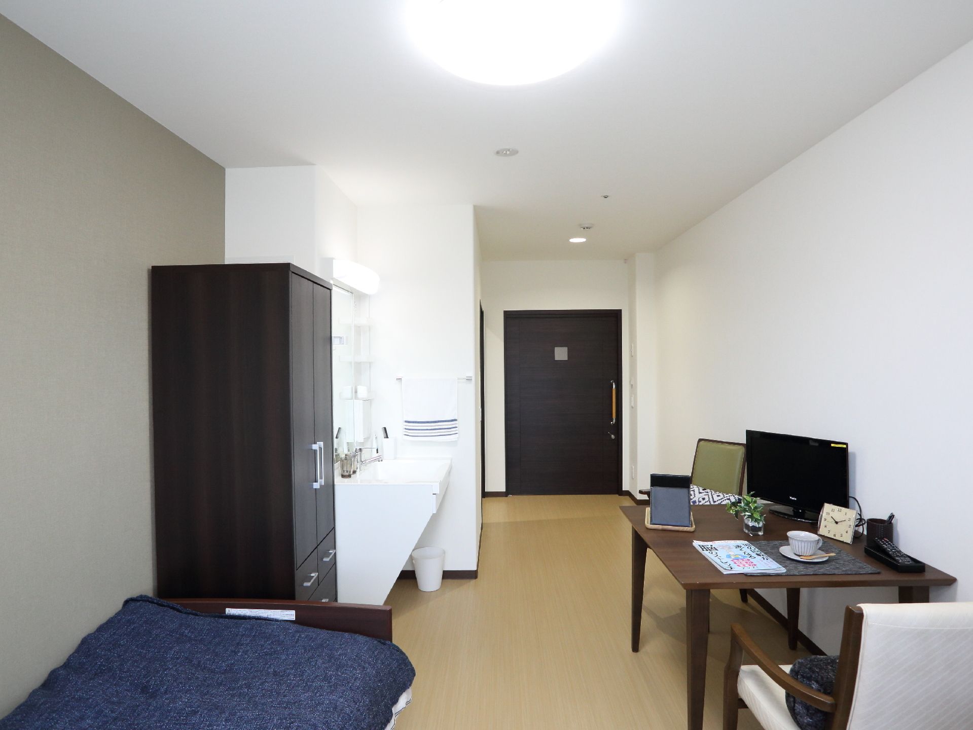 右側にはテーブルがあり、その上にはテレビや時計等が置かれている。左側にはベッドがある。正面奥には扉がある。