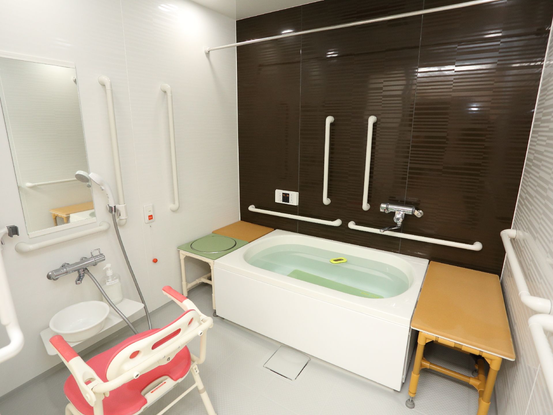 左側にはシャワーがあり、その手前には浴室用の介助椅子が置かれている。右側には湯船があり、その左右両側には台が置かれている。