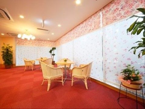 赤いカーペットが敷かれたエントランスには、机と椅子が置かれ、花や観葉植物が飾られている。大きくとられた窓に花柄のシェードが掛けられている。