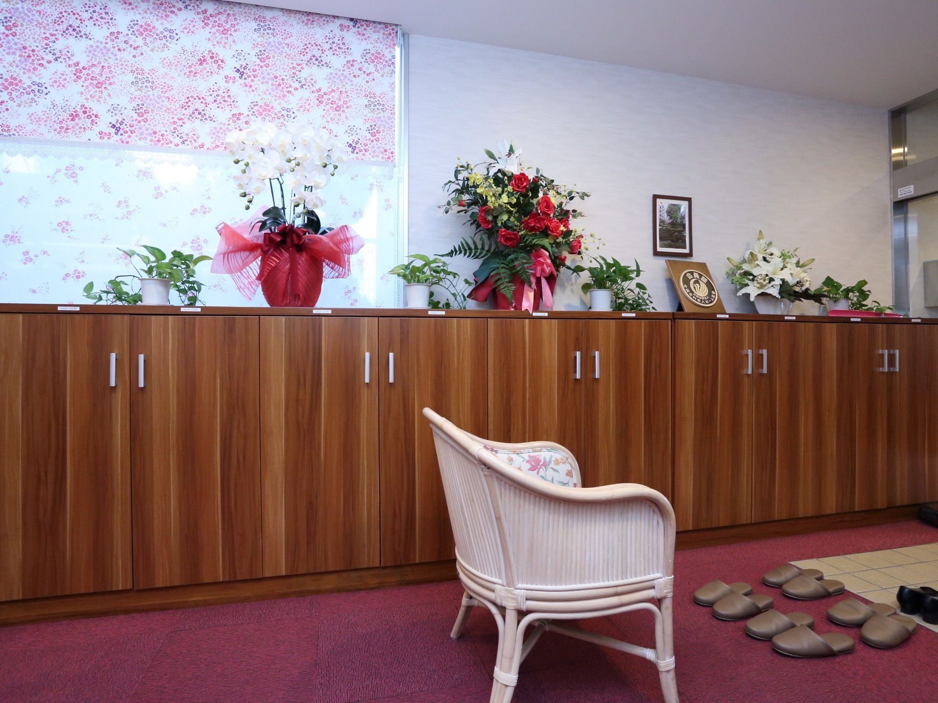 壁側には扉のついた大きな下駄箱がある。下駄箱の上には色鮮やかな花がいくつも飾られている。椅子が１脚置かれている。