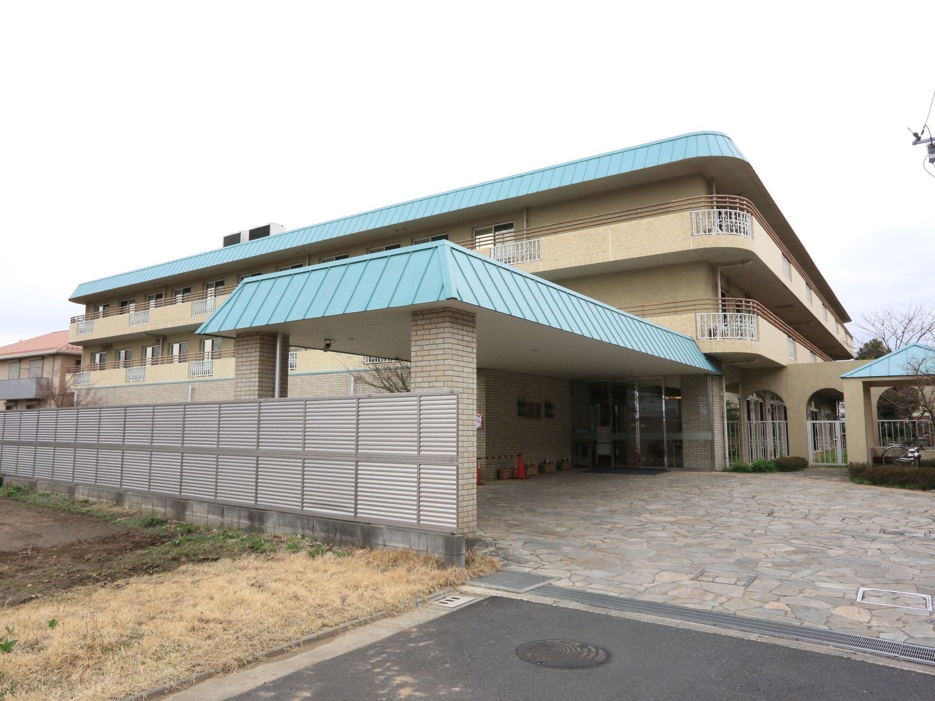 ベージュと水色のツートンカラーの３階建て施設の様子。周囲は住宅街となっている。２階と３階の各居室には大きなベランダが備わっている。