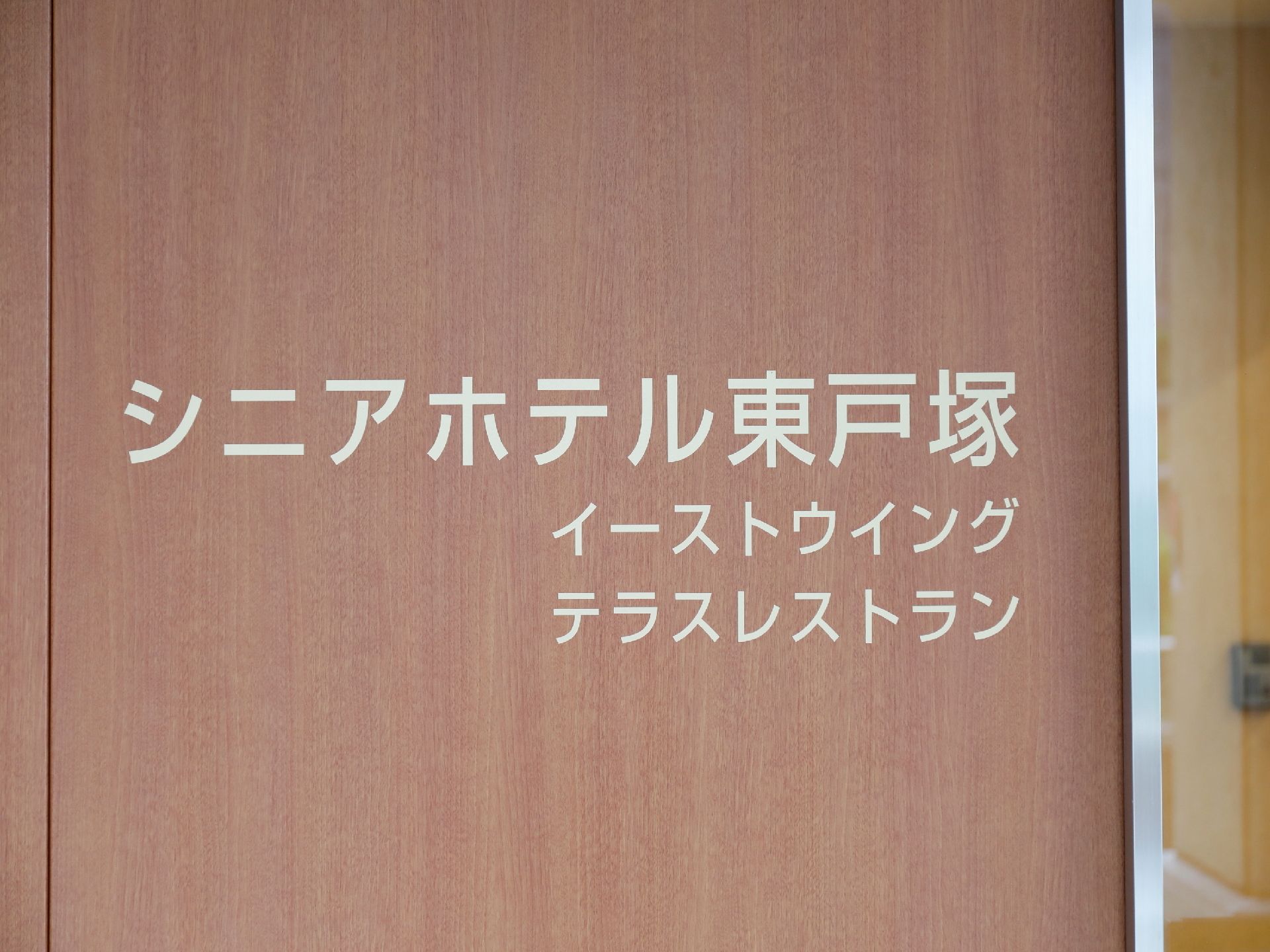 玄関ドアに表示された施設名。やさしい木目のドアにオフホワイトの文字で見やすい表示。テラスレストランの存在も表示されている。