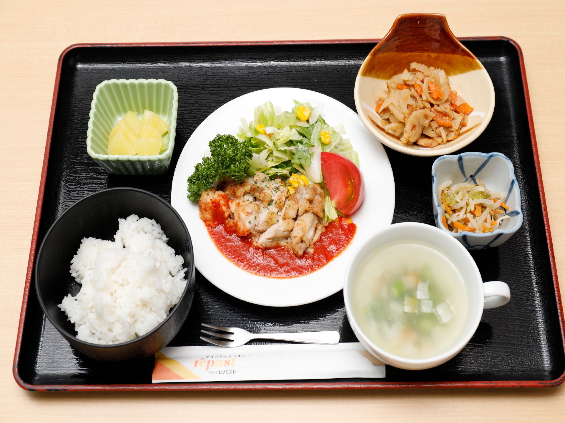 黒のトレーにご飯と汁物、真ん中にメインのおかずと3種類の小鉢の食事。手前に割りばしとホークが置かれている。