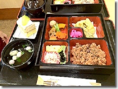 黒いトレーの上に置かれた食事。色とりどりのおかずが盛られた弁当箱の横に、汁物やケーキが置かれている。