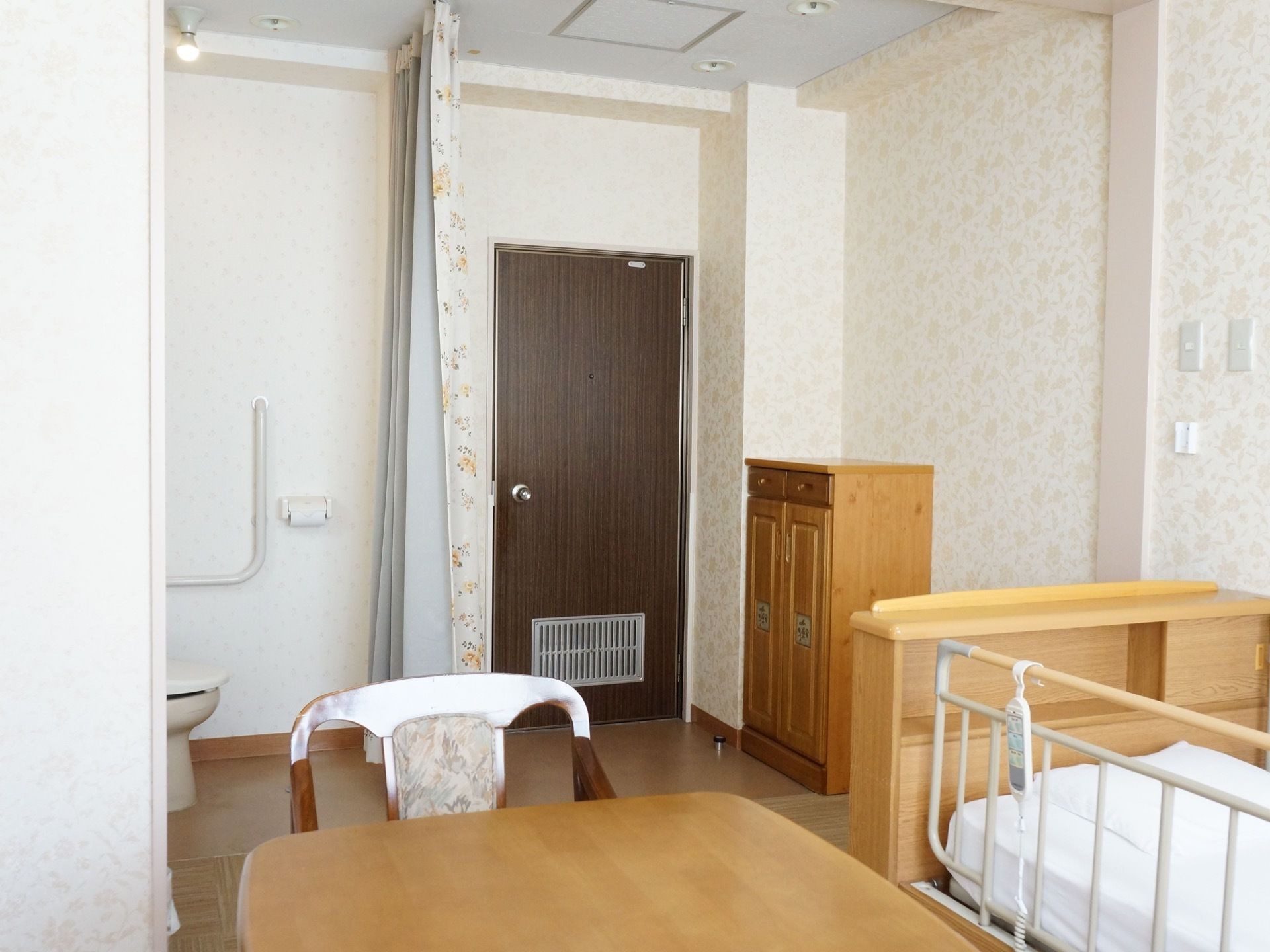 居室入口ドア側の様子。ドアをはじめ、電動ベッドや収納など木目調の家具が置かれている。居室内にトイレがある。
