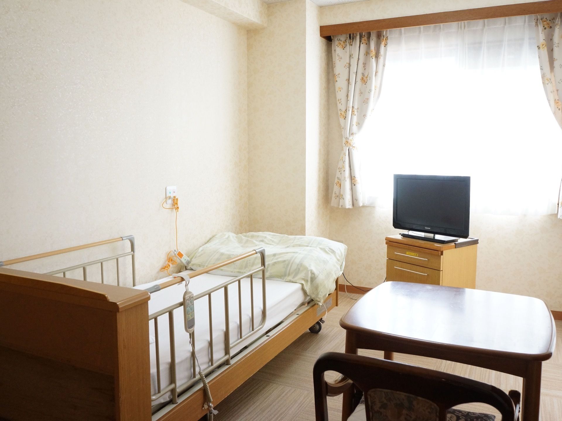 居室窓側の様子。電動ベッドとテレビ、椅子とテーブルが置かれている。壁面からナースコールがのびている。