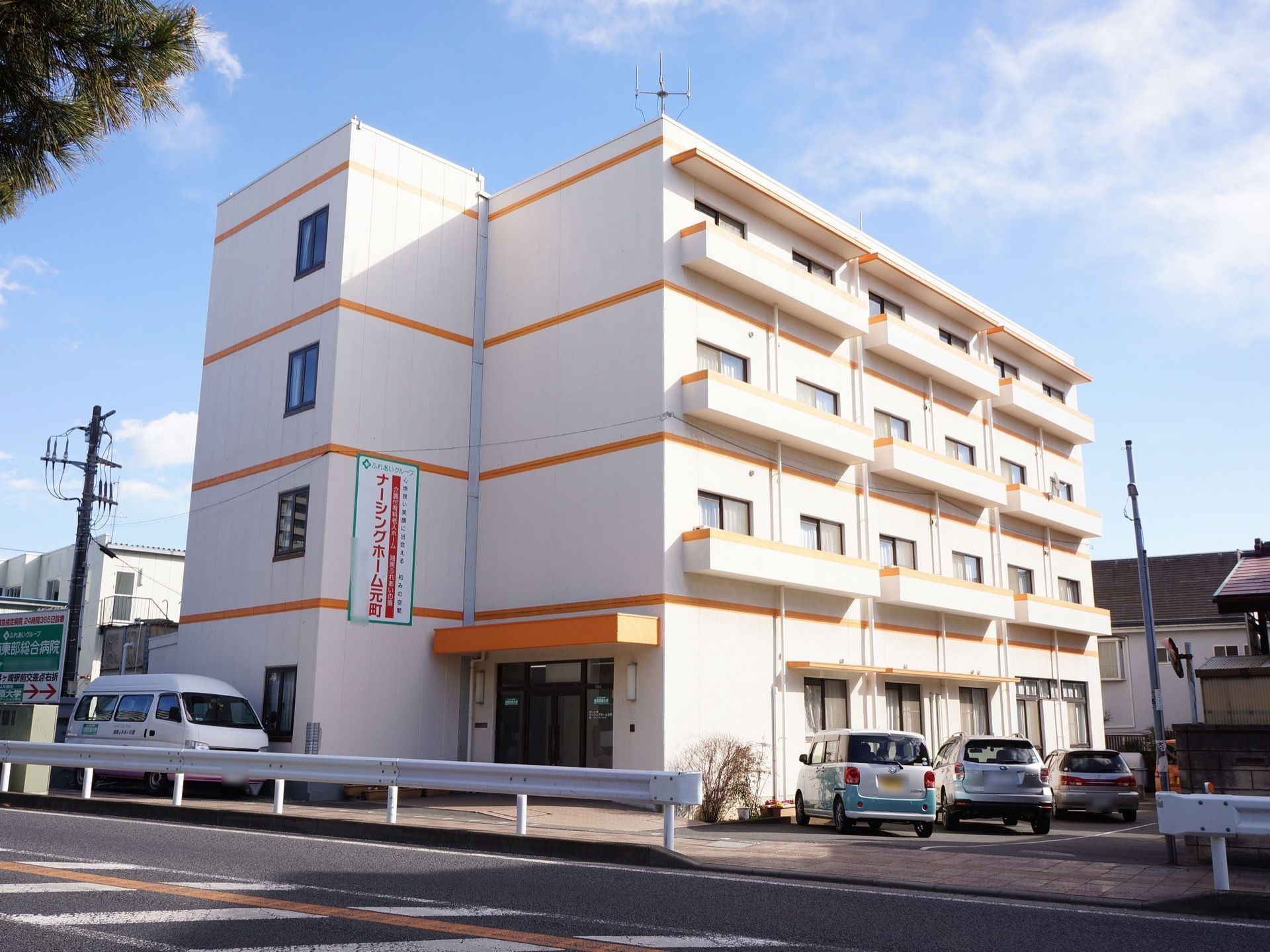 ４階建ての建物。ベージュとオレンジ色の外壁で、施設名の看板がかかっている。駐車場に車がとまっている。