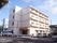 ４階建ての建物。ベージュとオレンジ色の外壁で、施設名の看板がかかっている。駐車場に車がとまっている。