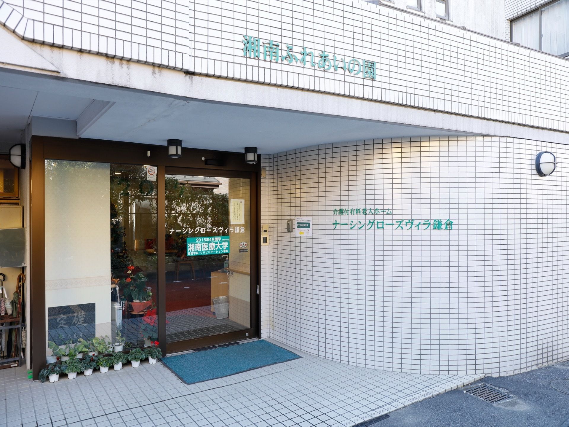 壁と玄関のドアに施設名が表記されている。扉は自動ドア。植木鉢が室内外に置かれている。傘立ても近くに置かれている。