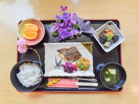 米飯、汁物、煮物、焼き魚、デザートの食事。食事トレーのうえに、折り鶴や、季節の花などで飾られている。