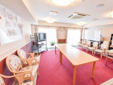 談話室の壁には、利用者の作った作品が飾られています。テーブルや椅子はもちろん、大きなテレビやホワイトボードが置かれている。
