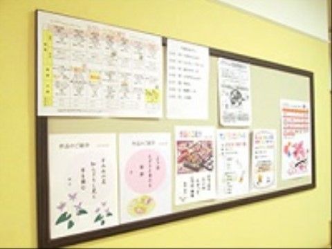 黄色い壁に、茶色い木枠の付いた掲示板が貼ってある。横長の掲示板には、A4やB4サイズのカラー印刷のプリントがある。