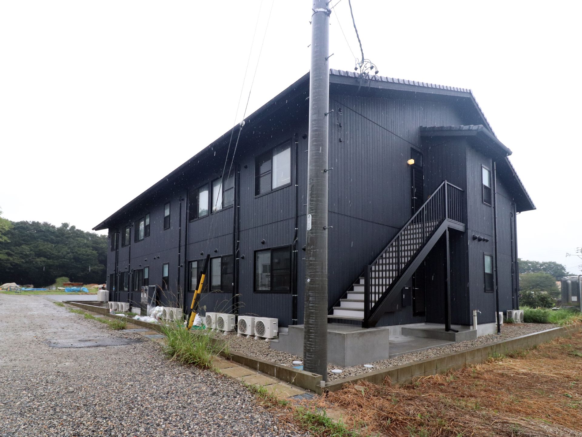 黒色の外観の建物で、上下に窓が並んでいる。横側には階段が設置してある。角には電柱があり、周辺は緑豊かな環境である。