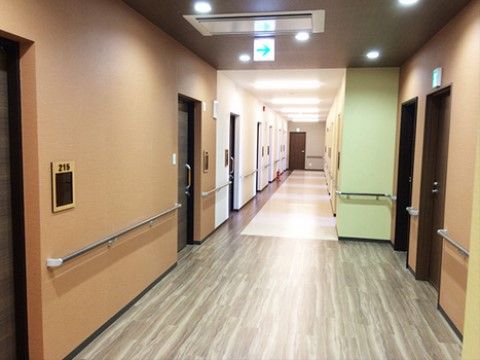 車椅子をご利用の方もスムーズに通れる広い廊下。淡い黄緑やピンクなど、場所によって壁の色が違っている。居室入り口は引き戸である。