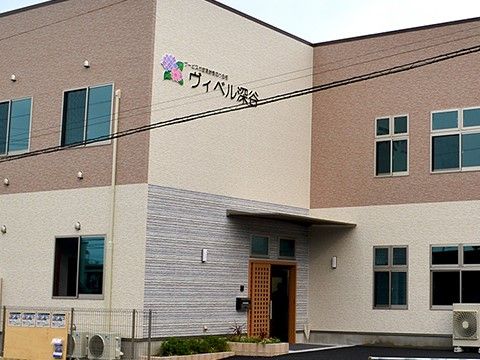 施設の建物の入り口付近の外観である。建物は2階建てになっており、壁はベージュ基調の落ち着いた配色である。